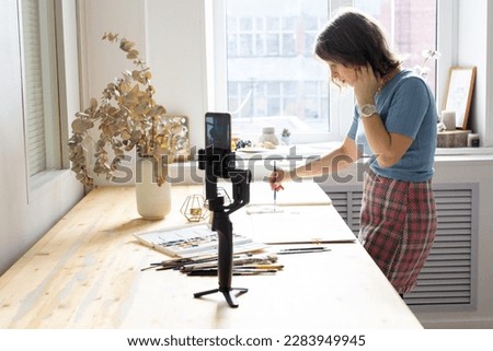 Young woman painting a picture with watercolor at wooden table, aquarelle process, girl sitting at the table and create picture, blogger filming video and making content