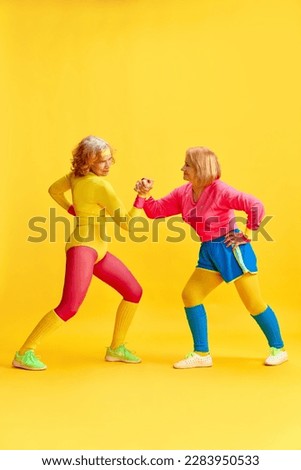 Arm wrestling. Two elderly sportive women in colorful uniform training, posing against yellow studio background. Concept of sportive lifestyle, retirement, health care, wellness. Ad