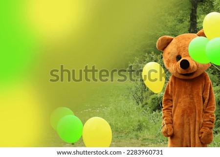 Man dressed as a teddy bear stands in a park surrounded by balloons. Colorful picture green yellow brown