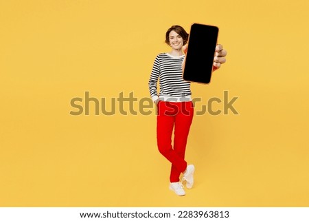 Full body young woman wear casual black and white shirt hold in hand use mobile cell phone with blank screen workspace area isolated on plain yellow color background studio portrait. Lifestyle concept