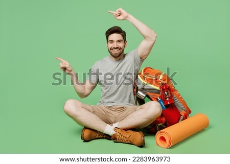 Full body young traveler white man near backpack mat sit point finger aside on area workspace isolated on plain green background. Tourist leads active lifestyle. Hiking trek rest travel trip concept Royalty-Free Stock Photo #2283963973