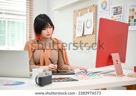 Attractive young Asian female web graphic designer or web editor working on her new design project, using graphic tablet and stylus, working in her studio.