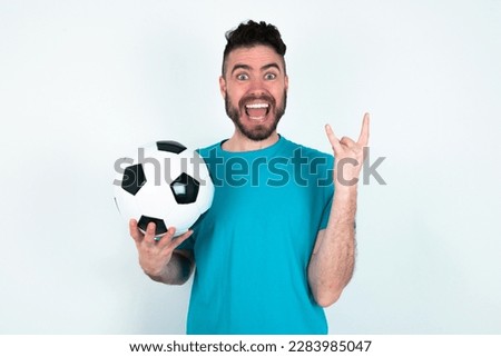 young handsome man wearing blue T-shirt over white background makes rock n roll sign looks self confident and cheerful enjoys cool music at party. Body language concept.