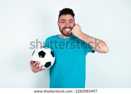 Happy young handsome man wearing blue T-shirt over white background  keeps fists on cheeks smiles broadly and has positive expression being in good mood