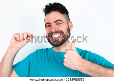 young handsome man wearing blue T-shirt over white background holding an invisible braces aligner and rising thumb up, recommending this new treatment. Dental healthcare concept.