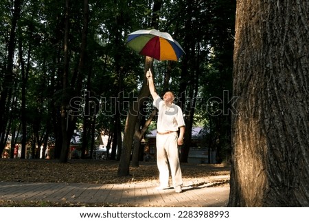 An elderly man stands under a large, multicolored umbrella. A pensioner on a walk in the park