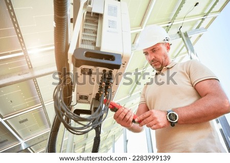Bottom view of worker checking solar inverter work. Measuring output voltage by current clamps. Adjusting inverter on back side of PV panel. Royalty-Free Stock Photo #2283999139