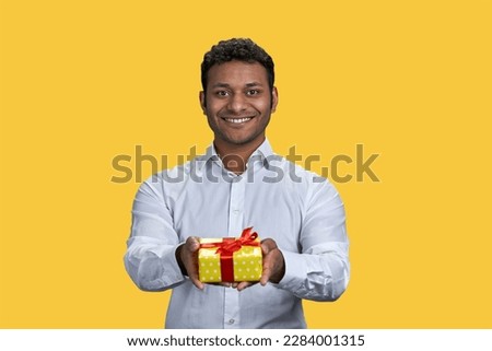 Portrait of young indian man giving a gift box. Take it concept. Greeting on holiday and sharing present. Isolated on yellow background.