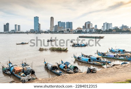 Small boats sit on muddy eastern bank of the river,much less developed than the riverside to the west.Setting sun reflecting on water,making silhouettes of small craft and distant city buildings.