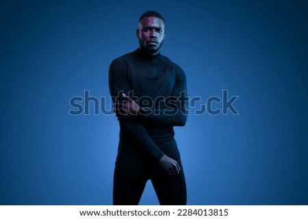 Thoughtful young brutal African American male model with dark hair and beard in total black activewear, with partial crossed arms looking at camera against blue background Royalty-Free Stock Photo #2284013815
