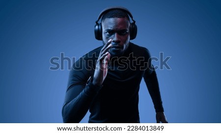 Determined African American sportsman in black turtleneck and earphones running on blue background in dark room while listening to music and looking at camera Royalty-Free Stock Photo #2284013849
