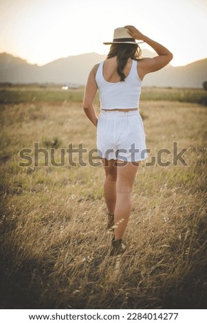  A beautiful young woman with long dark hair standing or sitting in a field of tall grass. The warm and gentle afternoon light enhances the natural beauty of the scene and creates a serene atmosphere.