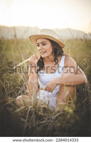  A beautiful young woman with long dark hair standing or sitting in a field of tall grass. The warm and gentle afternoon light enhances the natural beauty of the scene and creates a serene atmosphere.