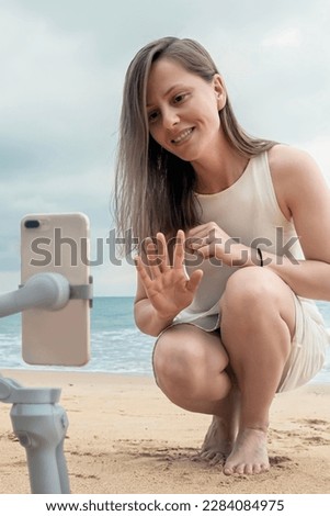 A young female travel blogger greets her viewers with her hand, taking pictures of herself in a blog on a smartphone on the beach by the sea, vertical photo
