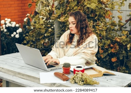 A young woman wrapped in a blanket works on a laptop at a table in the courtyard. There is a book and grapes on the table. The concept of a blogger or writer