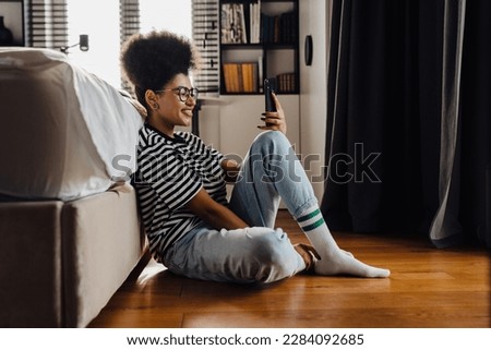 Young cheerful african woman wearing striped t-shirt using mobile phone while sitting on floor in bedroom Royalty-Free Stock Photo #2284092685
