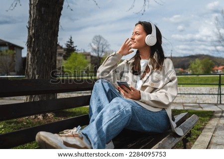 One young girl sitting on the bench in park listening to music on wireless headphones and using her phone Royalty-Free Stock Photo #2284095753
