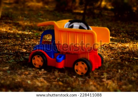 Colorful plastic toy dumpster truck with a hardhat in the back standing on the ground outdoors in the garden in the shade of a tree, side view