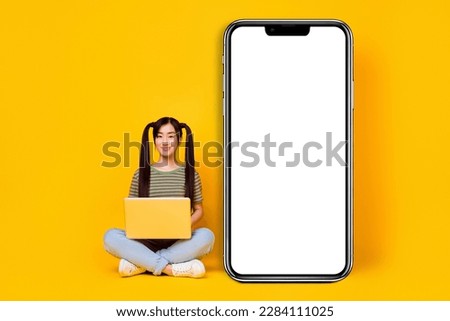 Photo of young girl working in laptop sit next to big phone display advertisement isolated on yellow color background