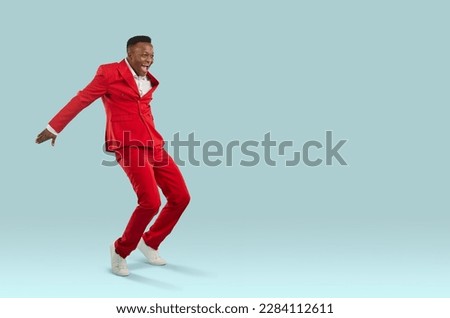 Happy African man in red suit having fun in fashion studio. Attractive ethnic guy in cool modern trendy outfit feeling elated and overjoyed dancing and jumping on light blue text copyspace background