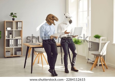 Animal people using laptop in office workplace. Team of 2 business men in unusual funny masquerade fantasy Halloween dinosaur horse masks working together, holding notebook device and leaning on desk