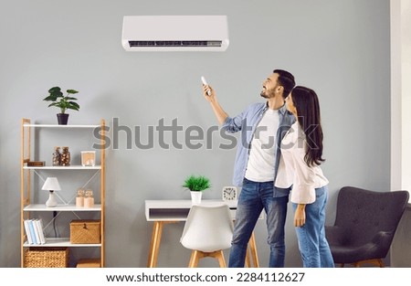 Young family couple using their modern air conditioning system at home. Happy husband and wife setting up the temperature on their white AC on the wall in the living room. Air conditioner concept Royalty-Free Stock Photo #2284112627