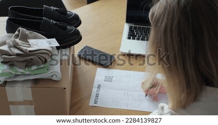 A young woman writes a return order and sits at a table with a laptop at home. Close-up of a female customer writing on paper and filling out a form, wanting to return a goods. Royalty-Free Stock Photo #2284139827