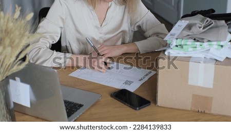 A young woman writes a return order and sits at a table with a laptop at home. Close-up of a female customer writing on paper and filling out a form, wanting to return a product. Royalty-Free Stock Photo #2284139833