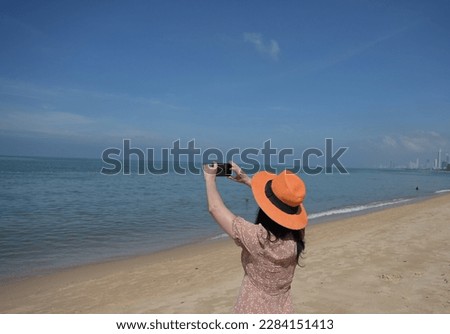 A adult female tourist Use both hands to lift up your mobile phone to capture the beauty of sea on a sunny day. A woman wearing light brown dress, an orange wide-brimmed hat is happy to take pictures.