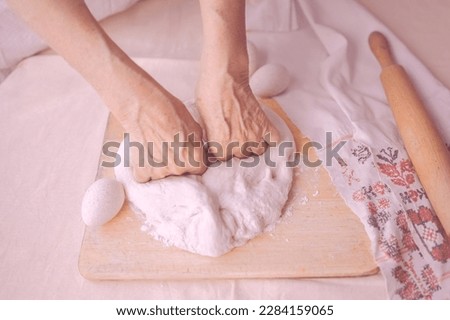 Overworked woman hands making pies out of dough. Elderly woman hands kneading dough, handmade homework with baked goods, eggs, cutting board and rolling pin, rustic embroidery towel.