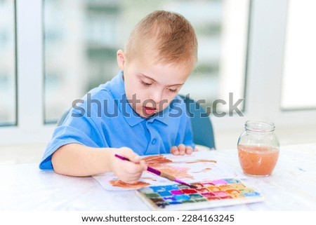 A cute boy with Down syndrome and autism draws a picture in an album with watercolors. Creative development of young children. The artist is learning the art of painting with paints.
