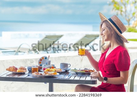 Young woman on summer vacation enjoying breakfast with tropical fruits, drinking orange juice on a luxury hotel resort terrace overlooking the sea.