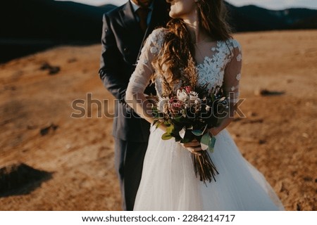 Bride and groom dressed elegant in their wedding gowns holding hands and hugging posing for their photo session in a desert like landscape by the lake. bride holding flower bouquet.