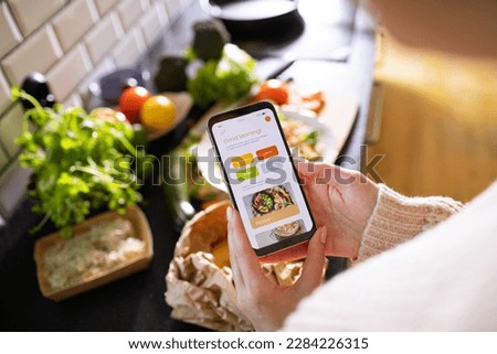 Young adult woman using a diet app on her phone while preparing a salad Royalty-Free Stock Photo #2284226315