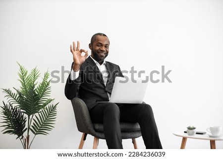 Diligent african employee in formal wear smiling sincerely at camera while typing on wireless laptop at office with modern minimalist design showing sign ok. People, work and technology concept.