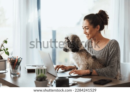 Attractive young female freelancer working on laptop from her home and having her pet dog in her lap to keep her company. Royalty-Free Stock Photo #2284241529