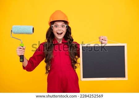 Little builder expert in helmet on construction site. Teen girl painter with painting brush tool or paint roller. Blackboard for copy space. Child on repairing work. Renovation concept.