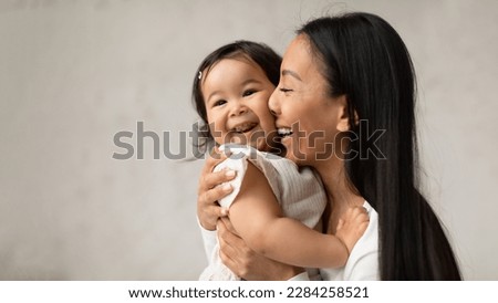 Mother's Love. Joyful Asian Mom Hugging Adorable Little Baby Daughter Rubbing Nose On Her Cheek Posing Over Gray Wall At Home. Mommy Cuddling With Infant. Happy Motherhood Moments. Panorama