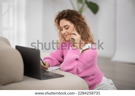 Young female freelancer working from home, using laptop and talking on cellphone with client while sitting on floor in living room, smiling woman having phone call and taking notes to notepad