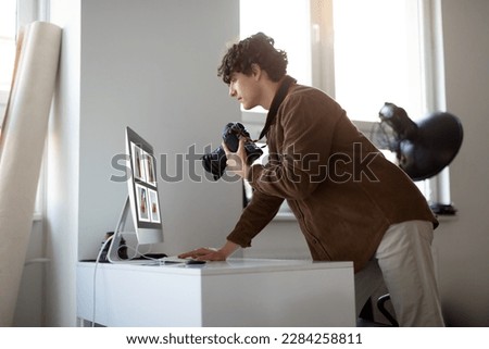Side view of professional male photographer holding camera and looking at computer monitor, working in photostudio. Man preparing retouching images. Creative agency.