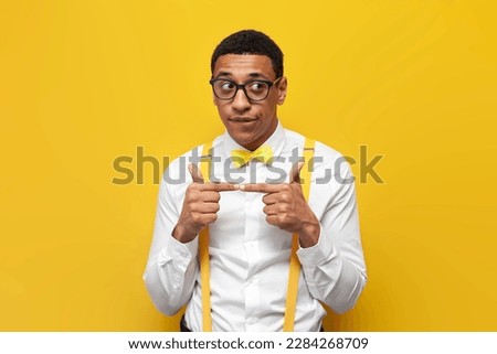 young insecure african american guy in white shirt with suspenders and bow tie is shy on yellow isolated background, nerdy man in glasses doubts