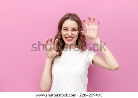 young cute woman in white t-shirt shows her claws with her fingers and imitates tiger, the girl growls and bares her teeth on pink isolated background Royalty-Free Stock Photo #2284269493
