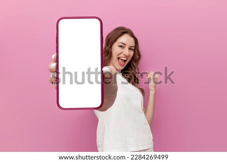 young cute woman in white t-shirt shows blank smartphone screen and rejoices in victory on pink isolated background, the winner girl advertises copy space on the phone, mockup Royalty-Free Stock Photo #2284269499