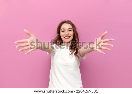 young cute woman in white t-shirt with outstretched arms wants hug, the girl hugs and holds her hands in front of her on pink isolated background Royalty-Free Stock Photo #2284269531