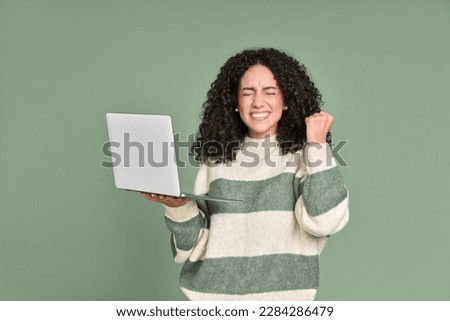Young happy latin woman winner holding laptop isolated on green background. Excited euphoric female model using computer winning online celebrating new great job offer with yes gesture concept.