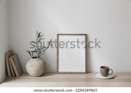 Mediterranean breakfast still life. Cup of coffee, books. Empty wooden picture frame mockup on desk, table. Textured vase with olive branches. Elegant working space, home office. Scandinavian interior