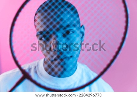 Male model looking at camera and holding badminton racket in front of his face. Sports with racket concept. Pink background studio shot. High quality photo