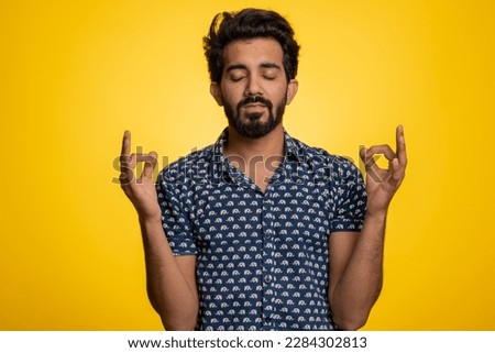 Keep calm down, relax, inner balance. Young indian man breathes deeply with mudra gesture, eyes closed, meditating with concentrated thoughts, peaceful mind. Hindu guy isolated on yellow background