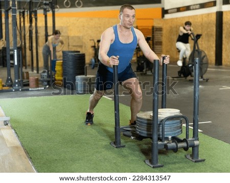 Portrait of concentrated strong adult man performing intense full-body strength training in gym, making effort to push heavy sled