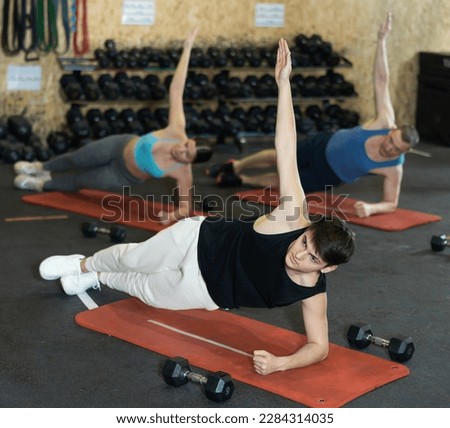 Concentrated young guy holding side plank pose to strengthen body muscles during intense group training in gym. Effective core exercises. Active lifestyle concept.. Royalty-Free Stock Photo #2284314035